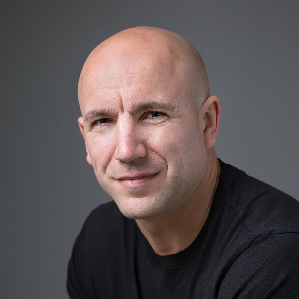 Close-up portrait of Andrew Kamphuis with a slight smile, wearing a black shirt against a gray background.