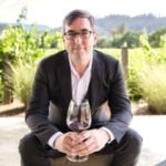 Man in suit holding a glass of red wine outdoors with vineyard background.