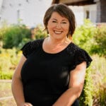 Smiling woman outdoors in garden with greenery and a house in the background.