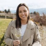 Smiling woman holding a glass of white wine outdoors with a scenic background.
